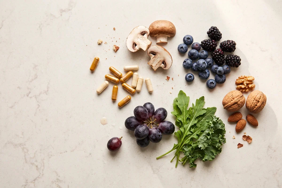 Assorted supplement capsules beside fresh fruit, vegetables, nuts and mushrooms on a bright kitchen counter.